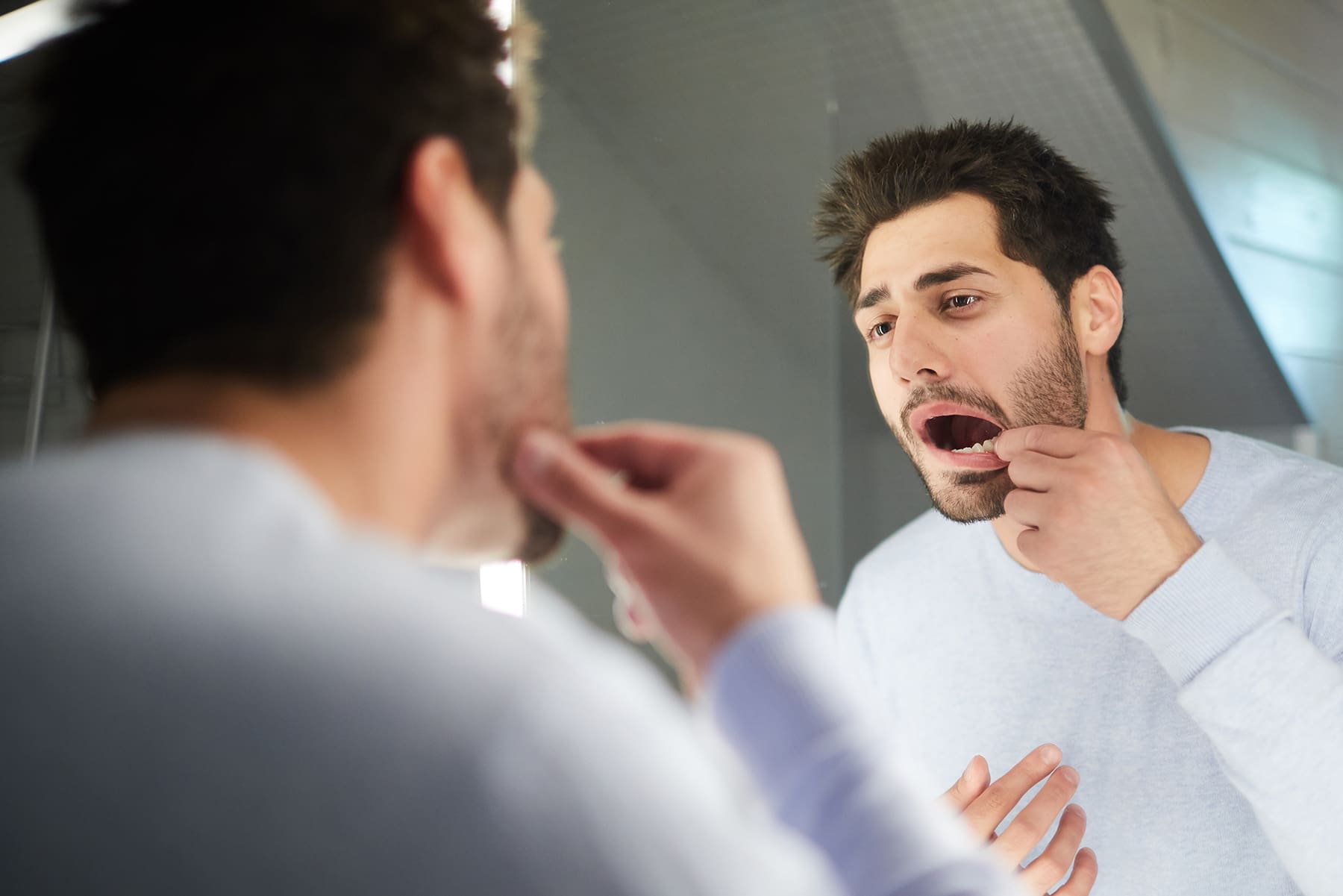 The image depicts a young man diligently flossing his teeth while looking at his reflection in a bathroom mirror, demonstrating proper technique and oral hygiene.